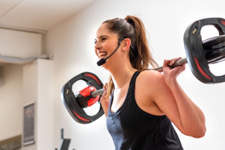 Group fitness instructor with a microphone headset holds a barbell on her shoulders