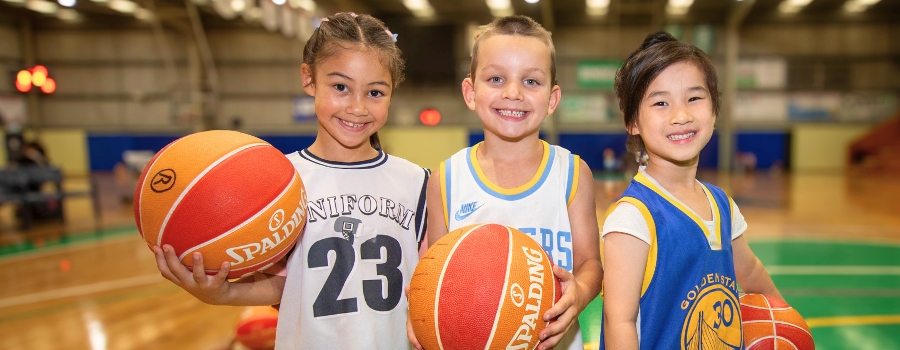 Three smiling children holding basketballs and standing on an indoor basketball court pose for a picture