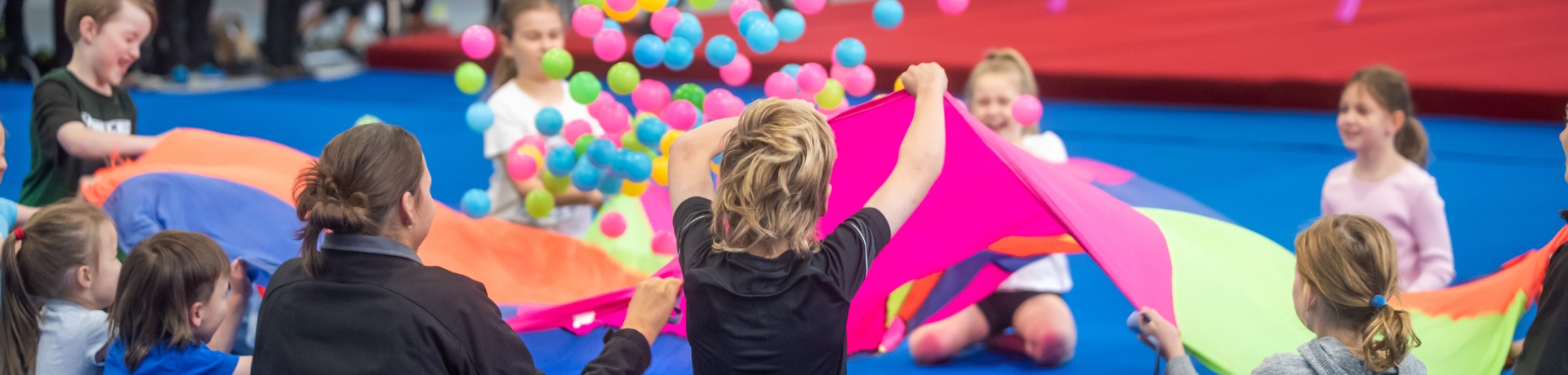 children in a gymnasium using a colourful parachute to bounce coloured balls into the air