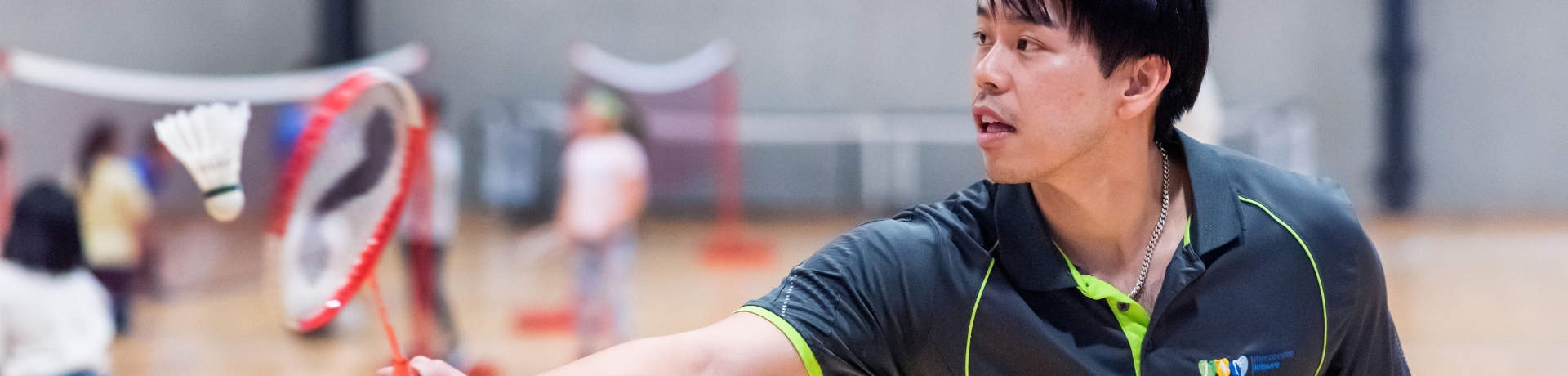 a coach demonstrates hitting a shuttle cock with a racquet while other children play badminton in the background